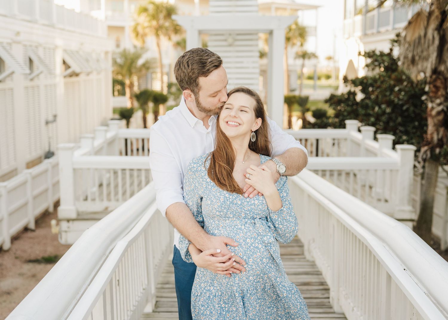 Expectant mother in a flowing blue floral dress stands on a wooden deck during golden hour.