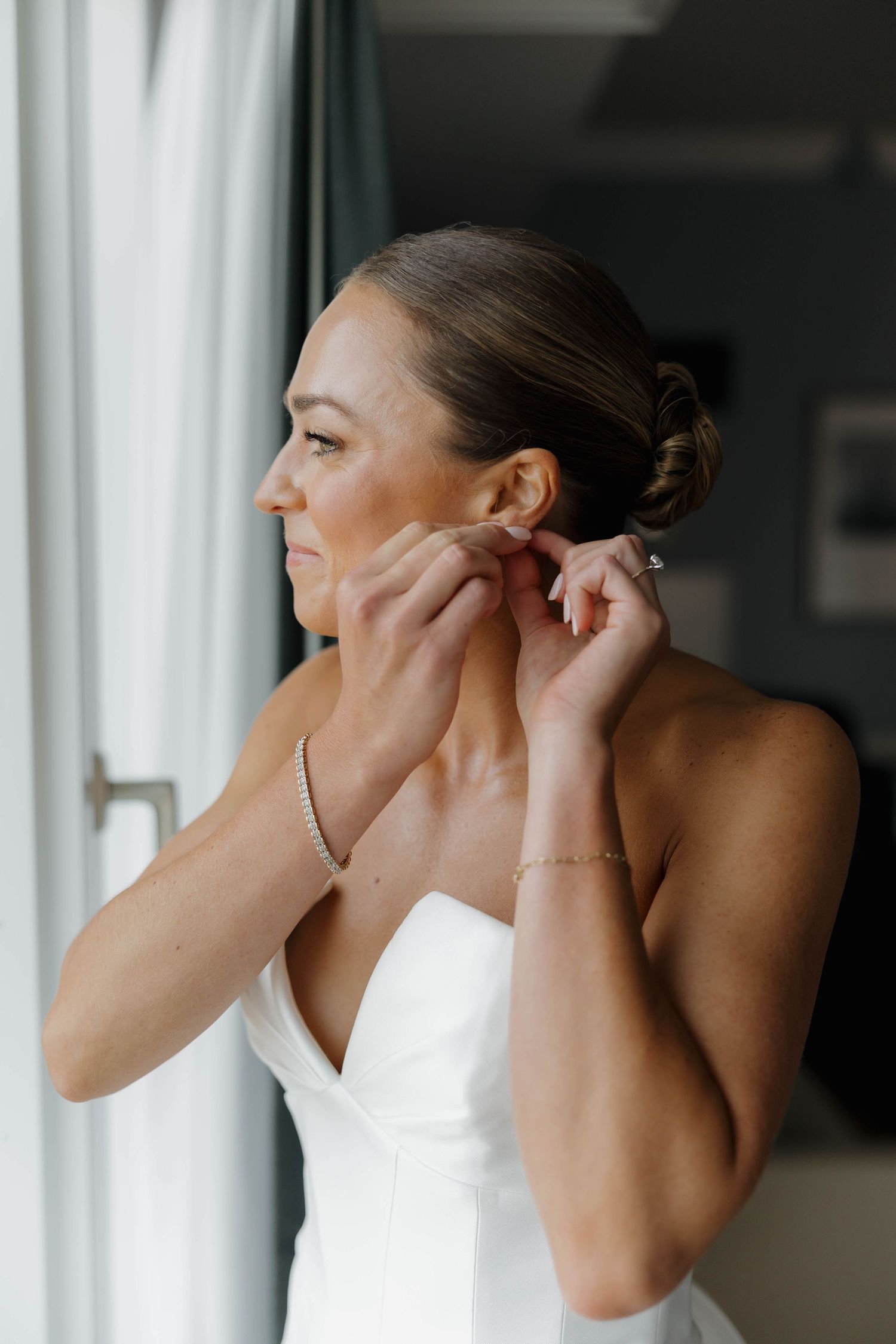 A person in a white dress puts on earrings while getting ready by a window.