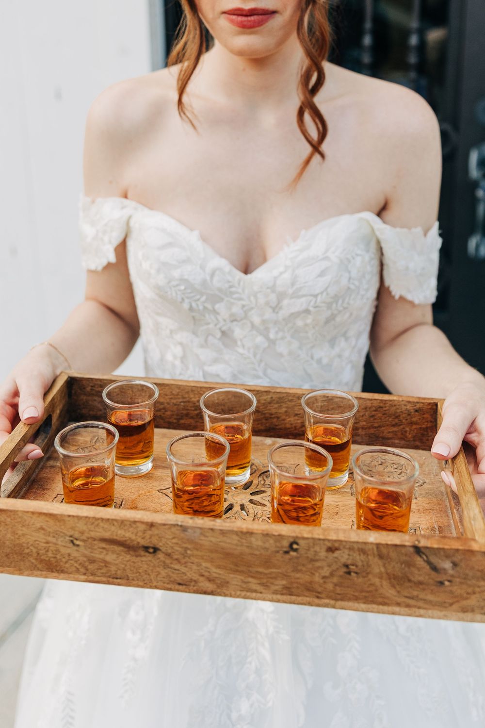 Three glasses of bourbon whiskey served on a rustic wooden tray with someone in a white dress holding it.