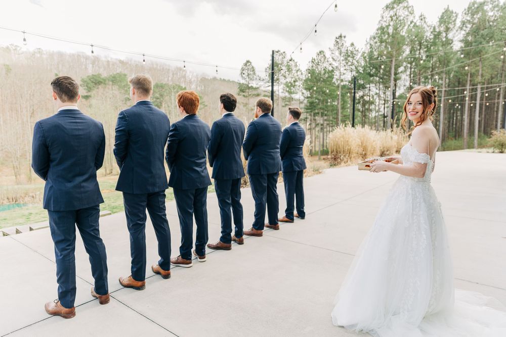 Wedding party in navy suits and white dress stand in line on rural road with trees in background.