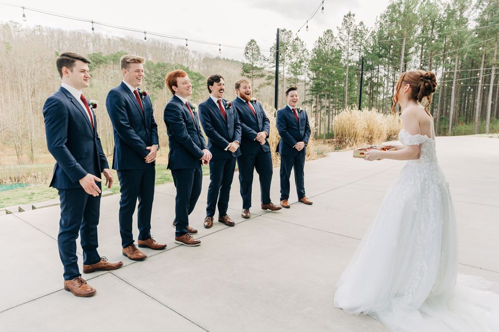 Wedding party stands in line against a wooded backdrop wearing navy suits and a white gown.