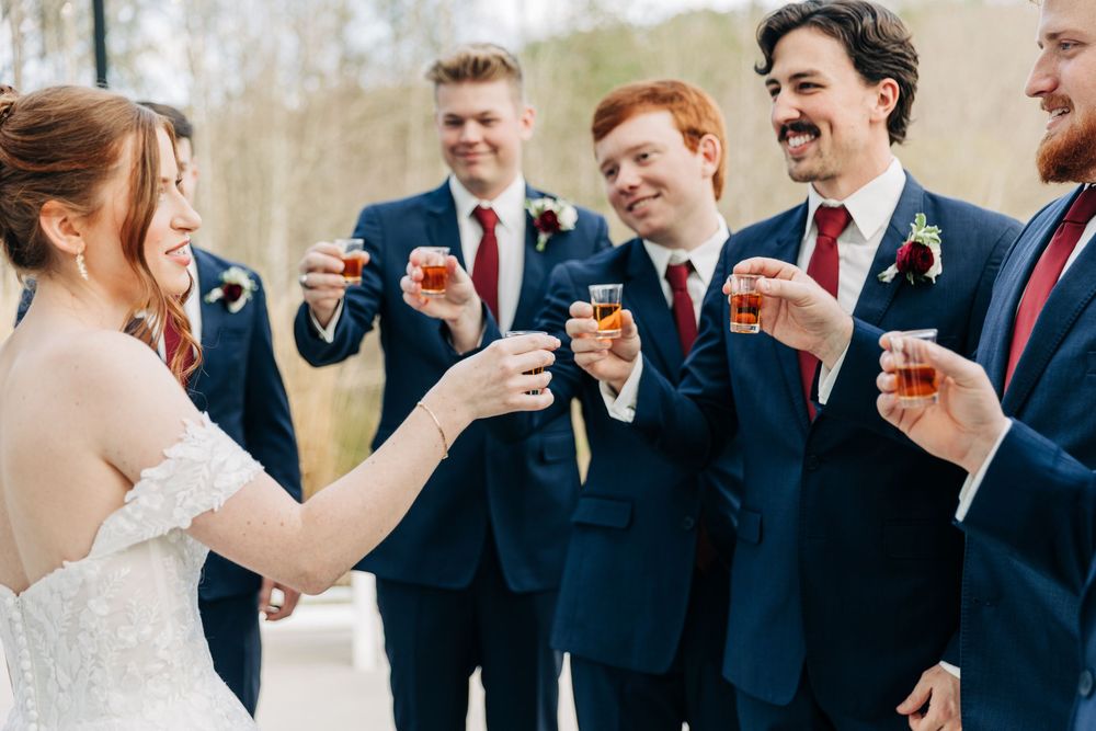 Wedding party in navy suits and a white bridal gown share laughs together during outdoor celebration.