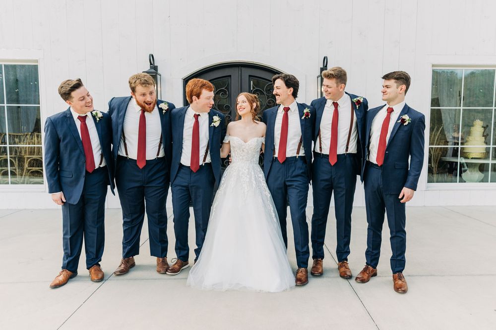 Wedding party in navy suits with red ties poses together on a bright outdoor terrace.