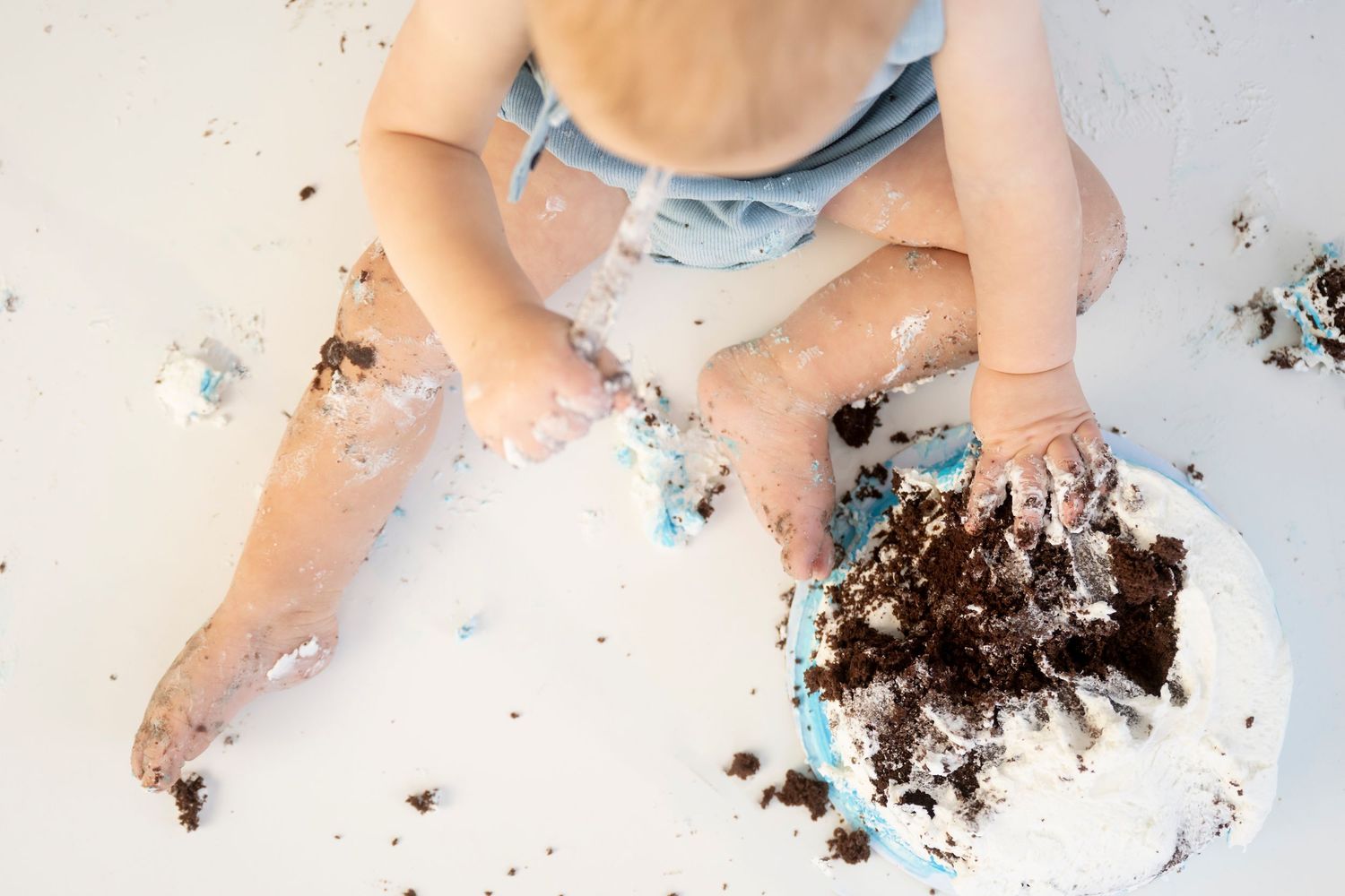A cake smash photo session with blue balloons and a baby in blue romper enjoying their first birthday cake.