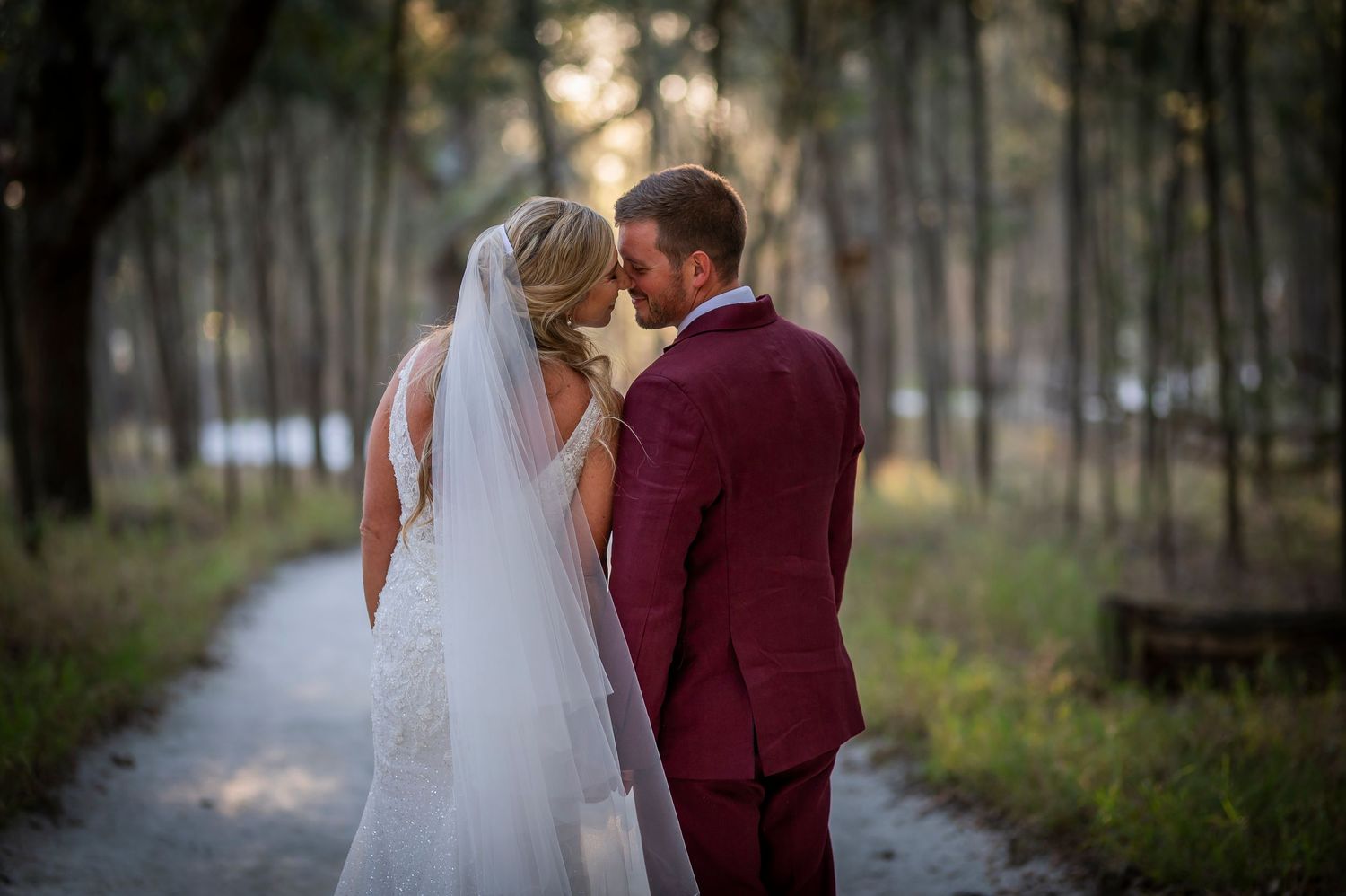Bride and groom share an intimate moment on a tree-lined path, she in a white gown with veil, he in a burgundy suit.