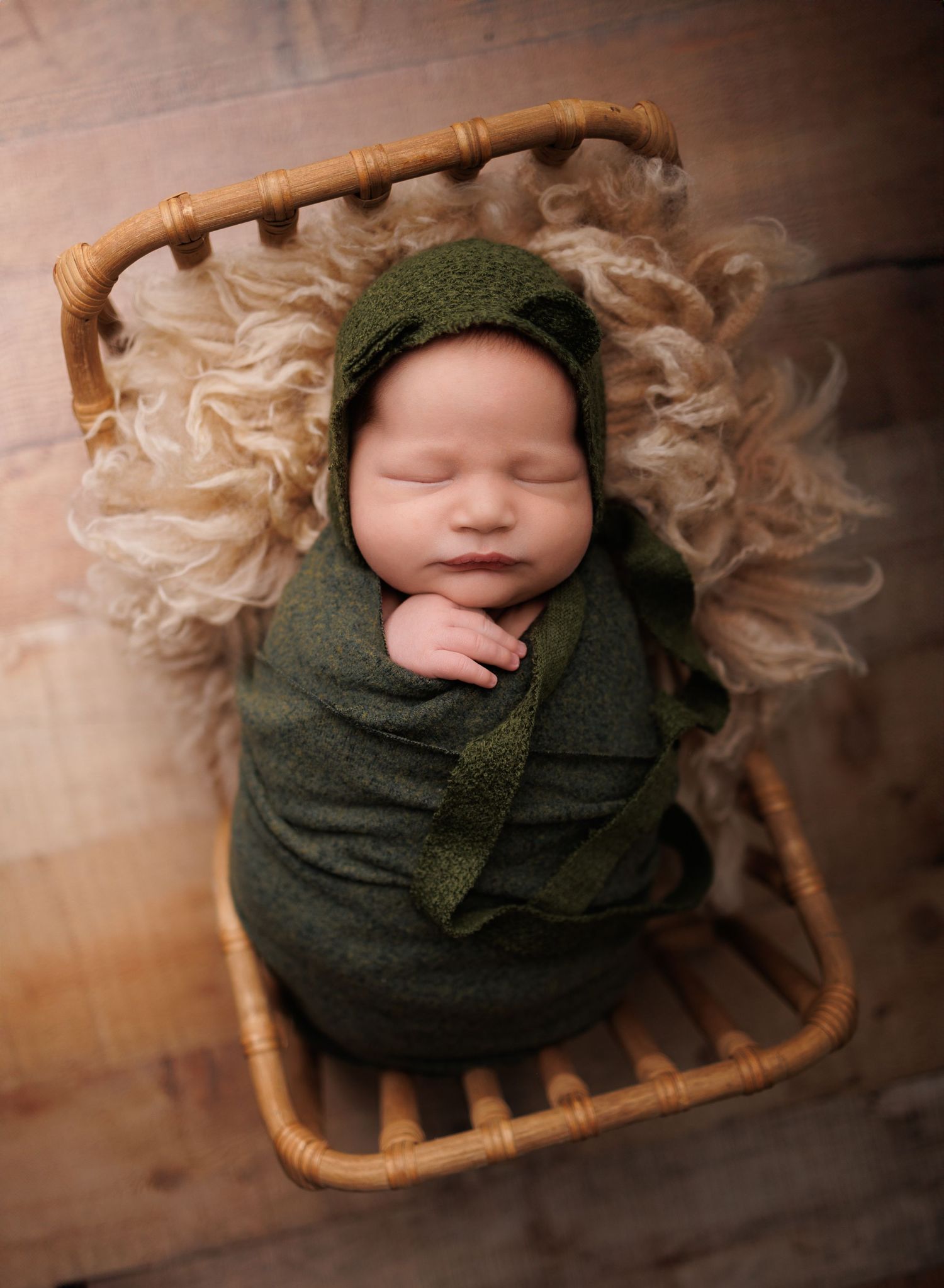 A photo of a sleeping newborn wrapped in a dark green blanket rests peacefully in a woven basket against a neutral backdrop.