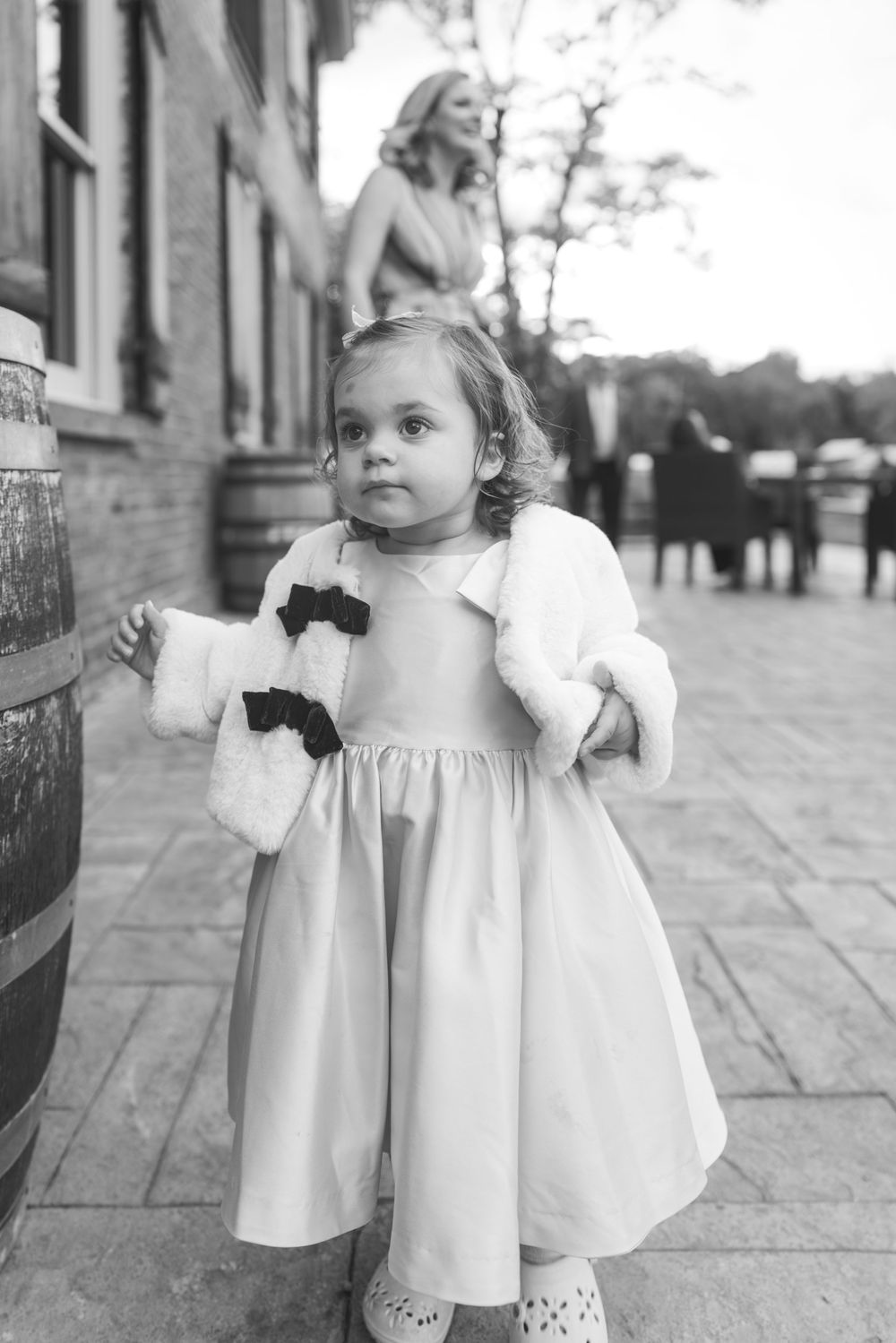 A young child in a white dress with black bows stands on a sidewalk in this vintage black and white photograph.
