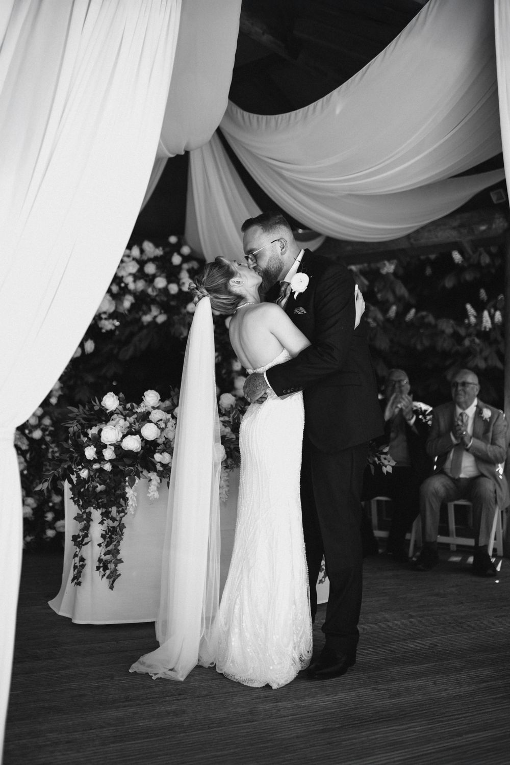 A couple shares their first kiss as newlyweds under elegant white draping at an indoor wedding ceremony.