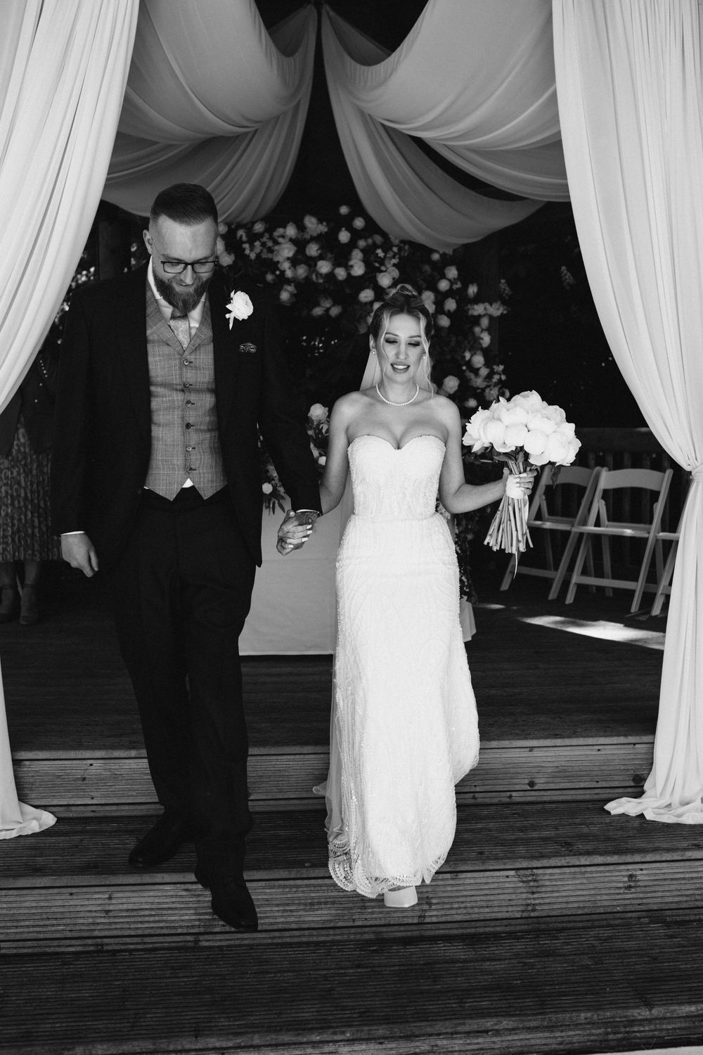 A black and white wedding photo shows a couple walking together under elegant white draped fabric at an indoor venue.