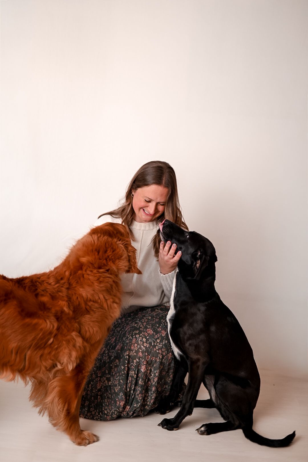 Woman sitting on floor smiling with a golden retriever and black Labrador dog in a bright studio.