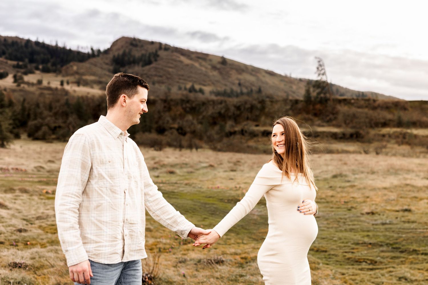 Pregnant couple holding hands in a field with rolling hills, wearing cream outfits during an outdoor maternity photoshoot.