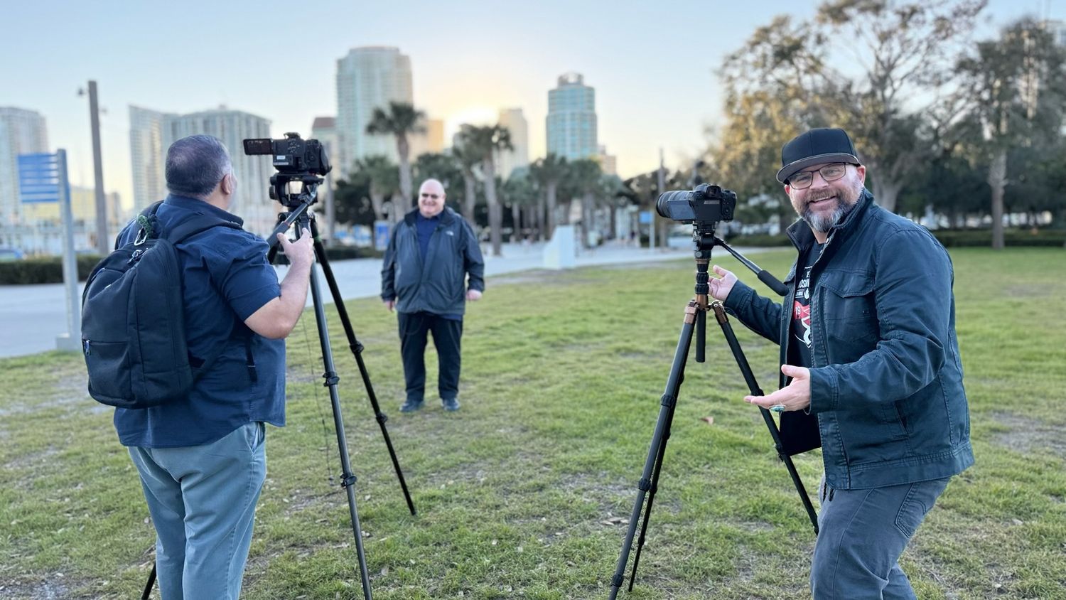 Three photographers with cameras on tripods filming outdoors in a park with city skyline at sunset.
