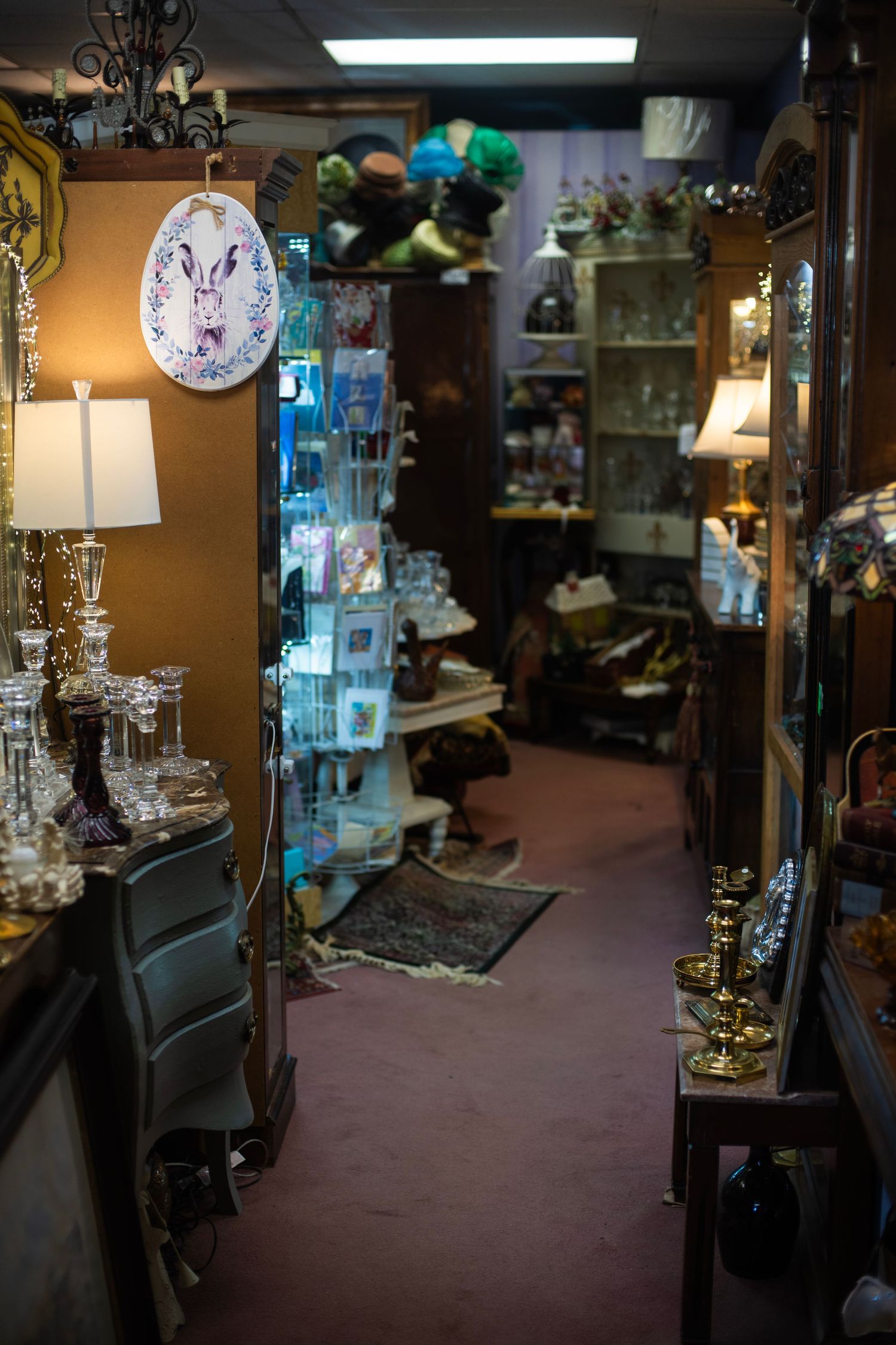 Cluttered antique shop aisle filled with lamps, glassware, candlesticks, and vintage decor items on wooden shelves.
