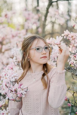 Young person in glasses and pink sweater poses among delicate spring magnolia blossoms.