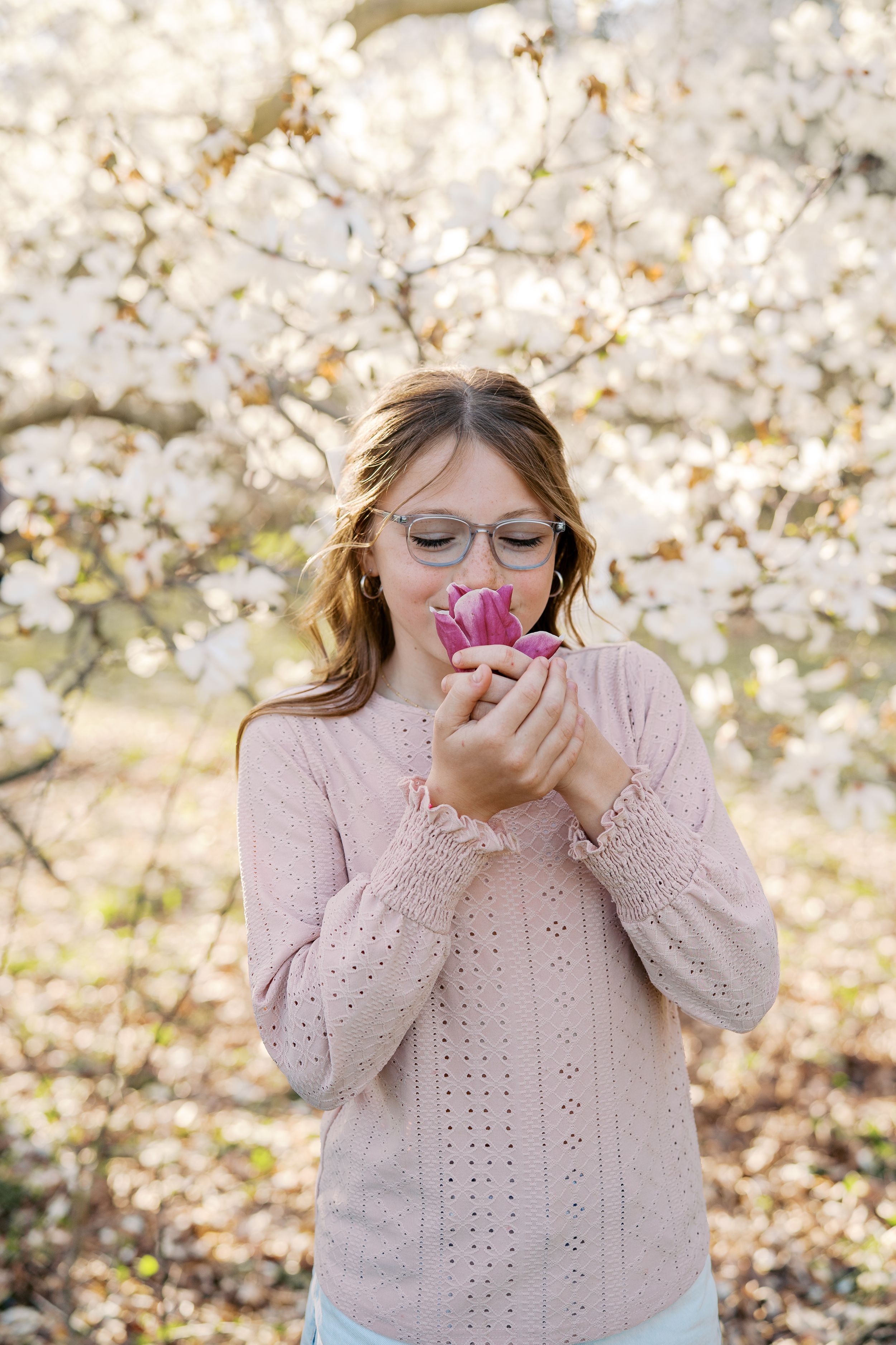 A person in a pink sweater sneezes among blooming cherry blossom trees in a spring garden setting.