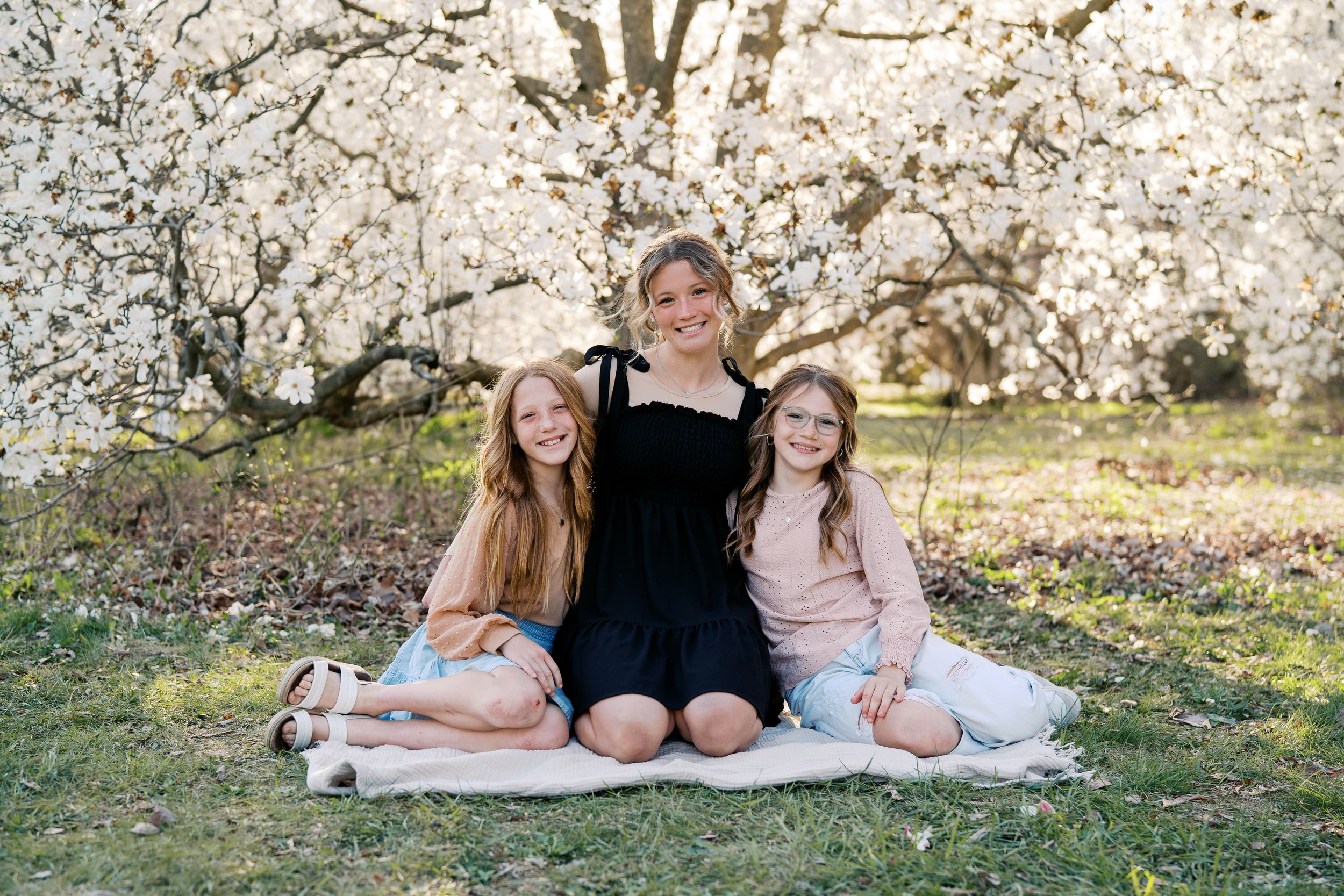 Three children sit on a blanket under blooming spring trees with white blossoms in a sunny garden setting.