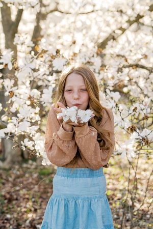 Person in blue dress and brown sweater playfully blowing cherry blossom petals in springtime.