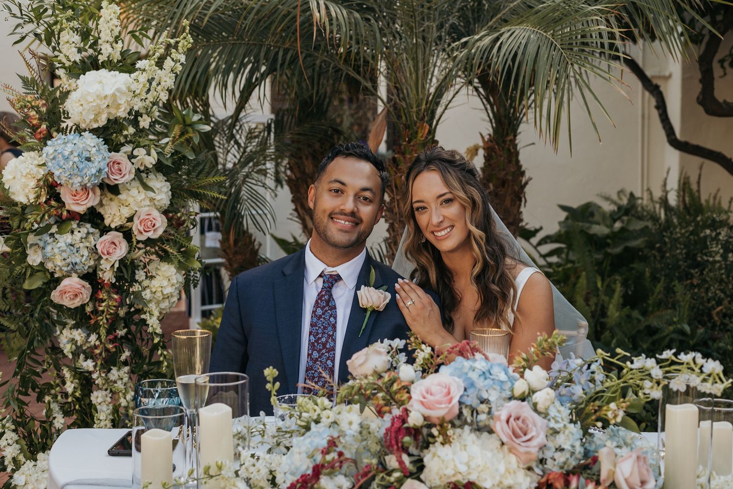Smiling bride and groom seated at floral wedding reception table surrounded by lush greenery and pink and blue flower arrangements.