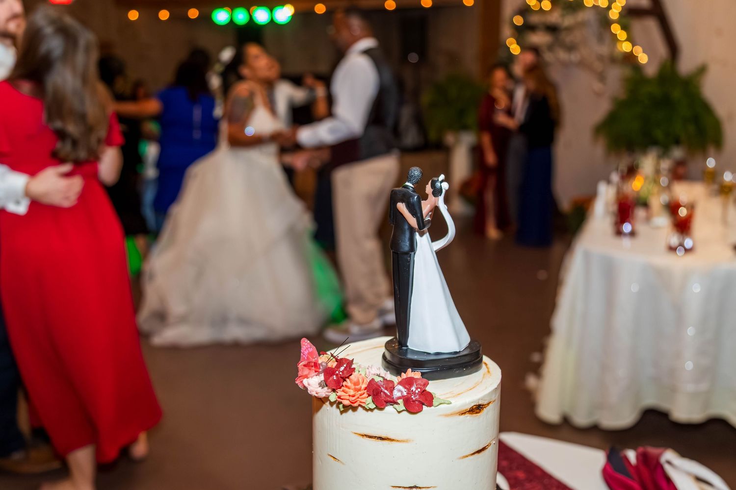 Wedding cake topper featuring bride and groom figurines atop a white cake, with guests dancing in the background.