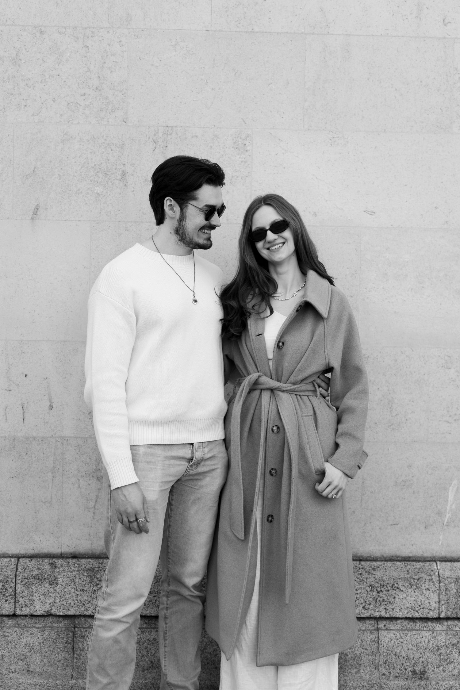 Stylish couple in sunglasses smiling together against a stone wall in a black and white portrait.