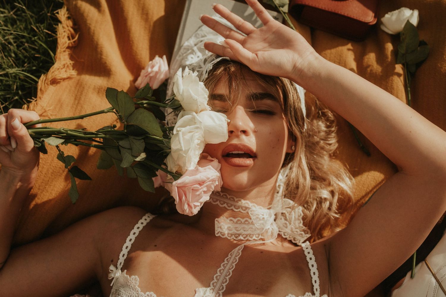 Woman lying outdoors holding white and pink roses, wearing lace top, with golden hour sunlight in a dreamy aesthetic.