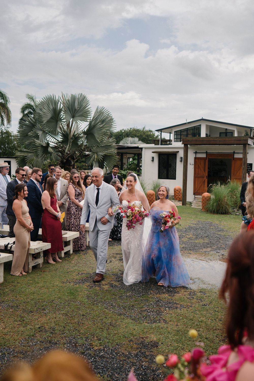 Wedding guests gather outside a modern venue with palm trees and gravel landscaping during an outdoor celebration.