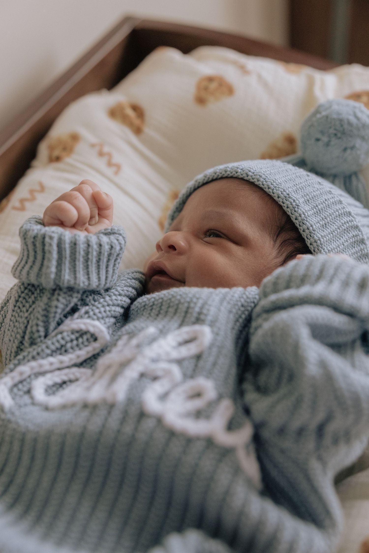 A newborn baby wearing a cozy blue knit sweater and matching hat rests peacefully on white bedding.