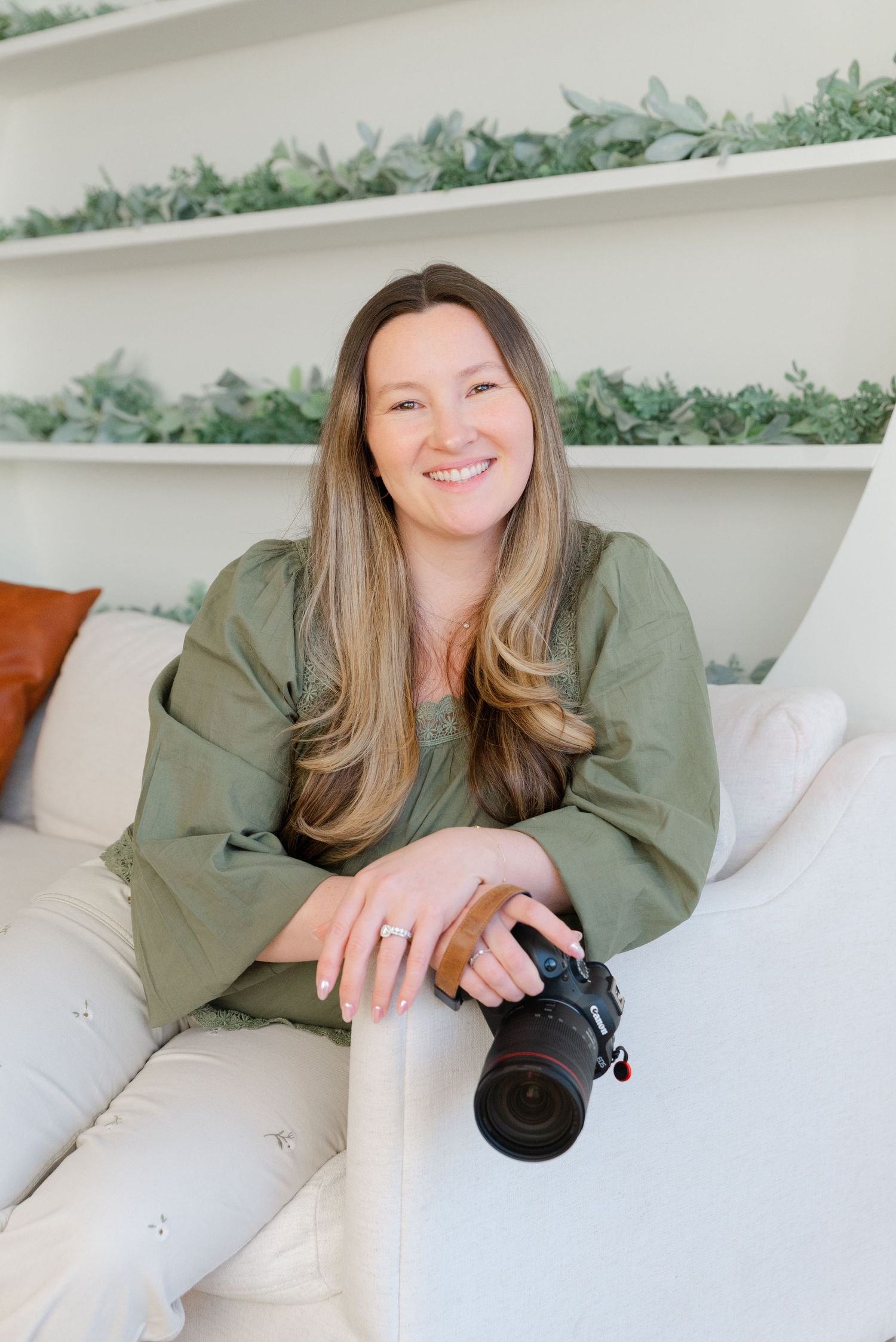 A person in an olive green top relaxes on a white couch with greenery decor visible in the background.