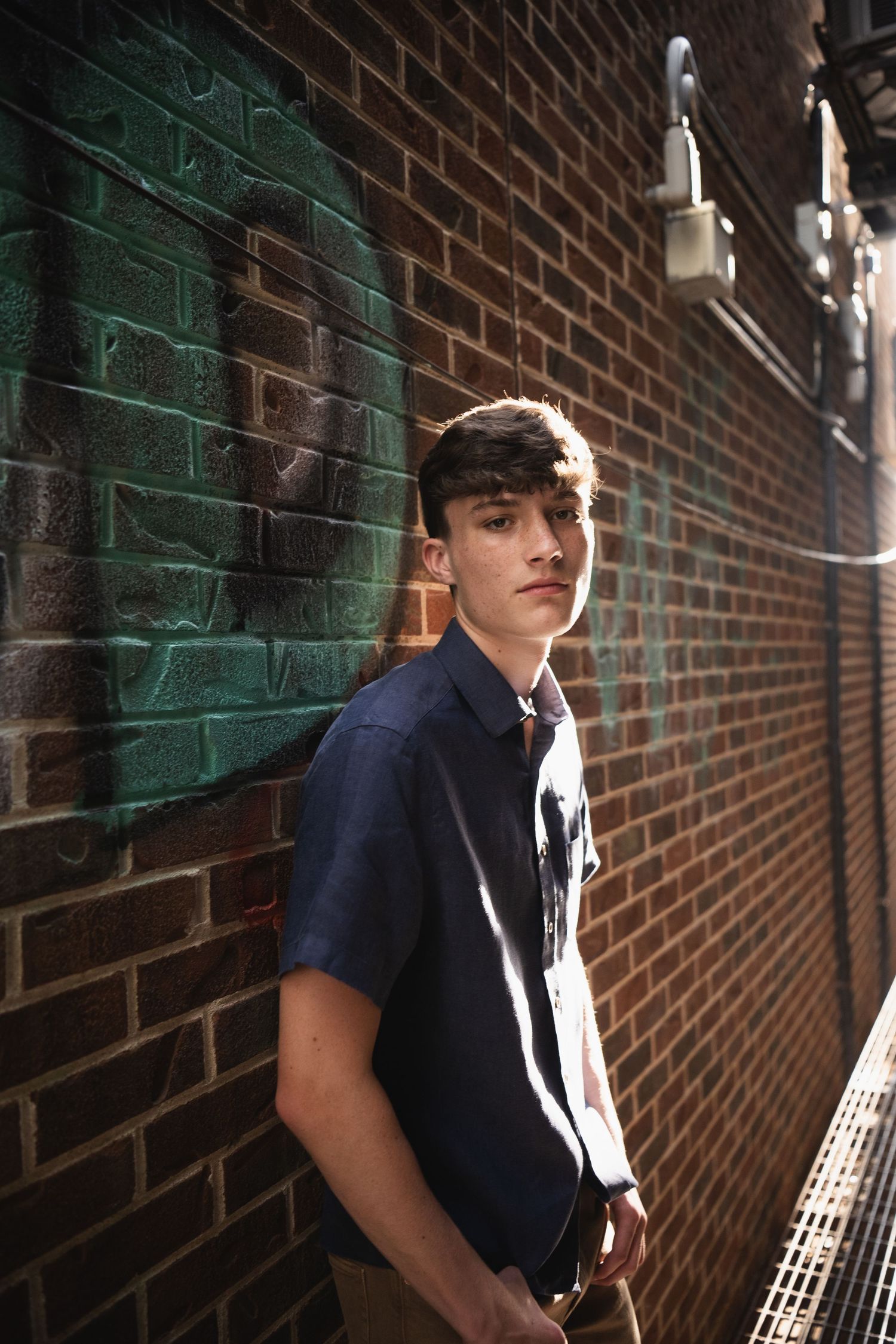 A young person in a navy blue shirt poses against a brick wall with green painted accents.