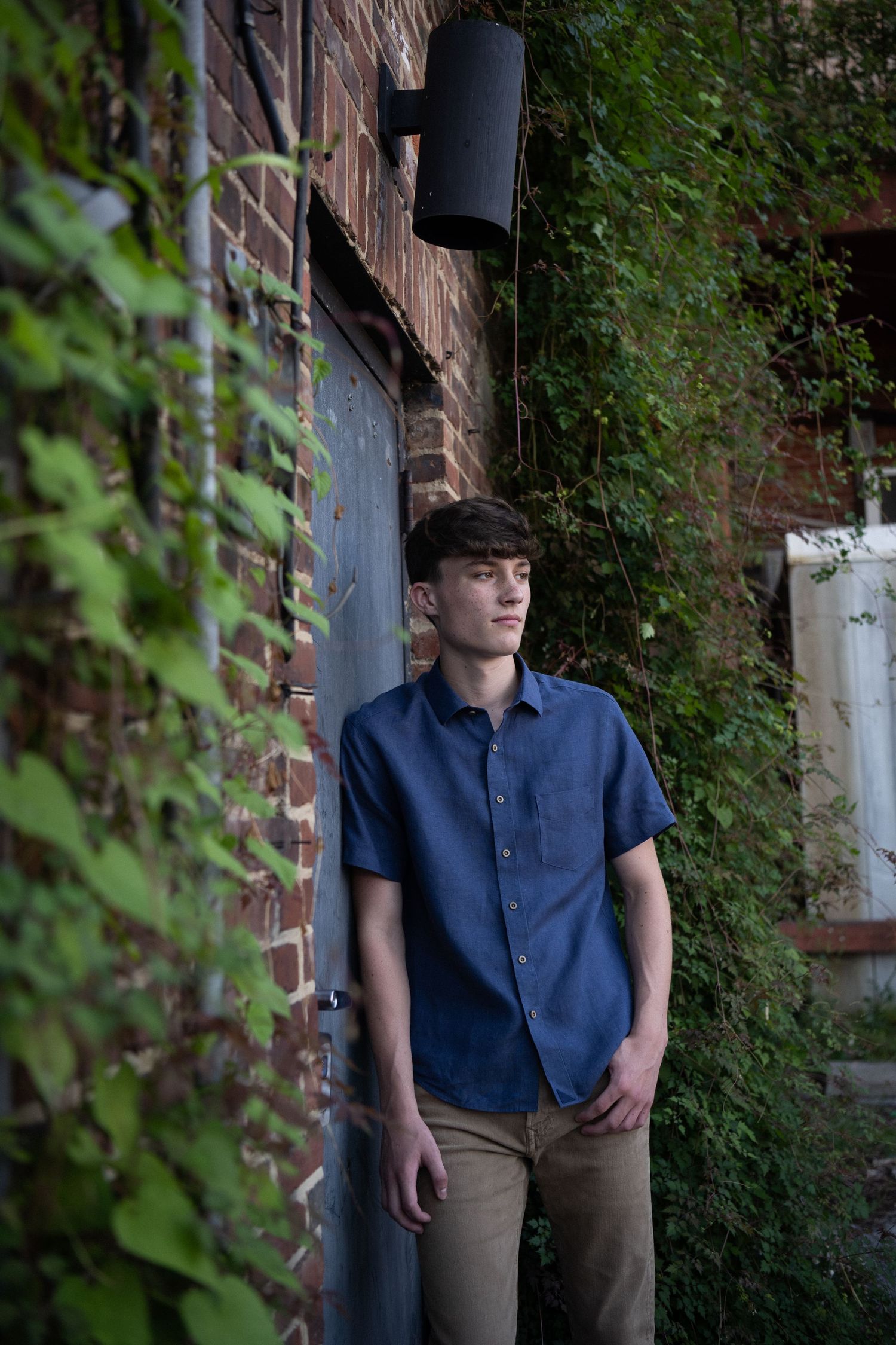 A person in a navy blue button-down shirt and khaki pants stands against a brick wall with ivy in an outdoor setting.