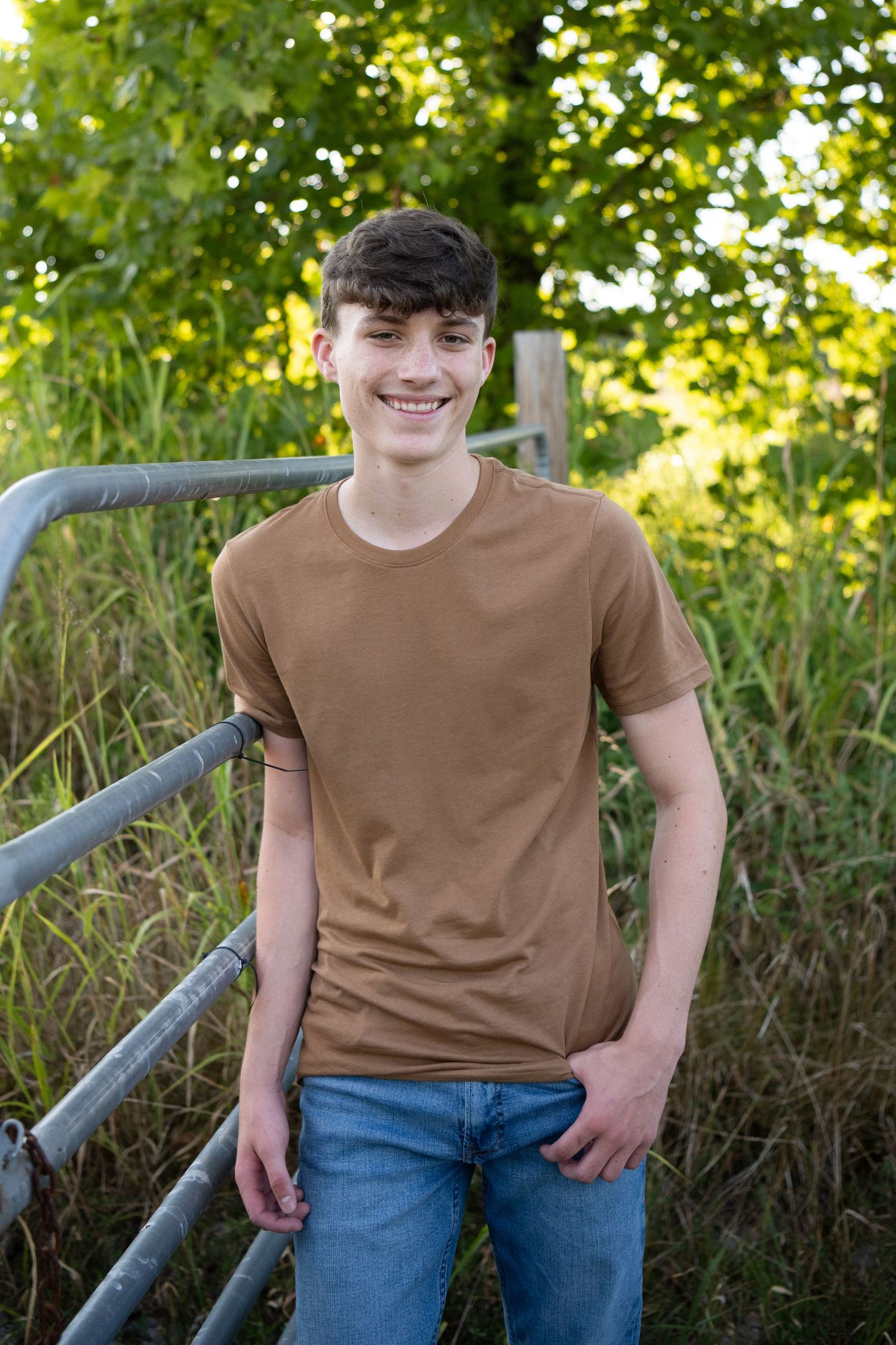 A person in a brown t-shirt and blue jeans poses outdoors against a green leafy background in natural lighting.
