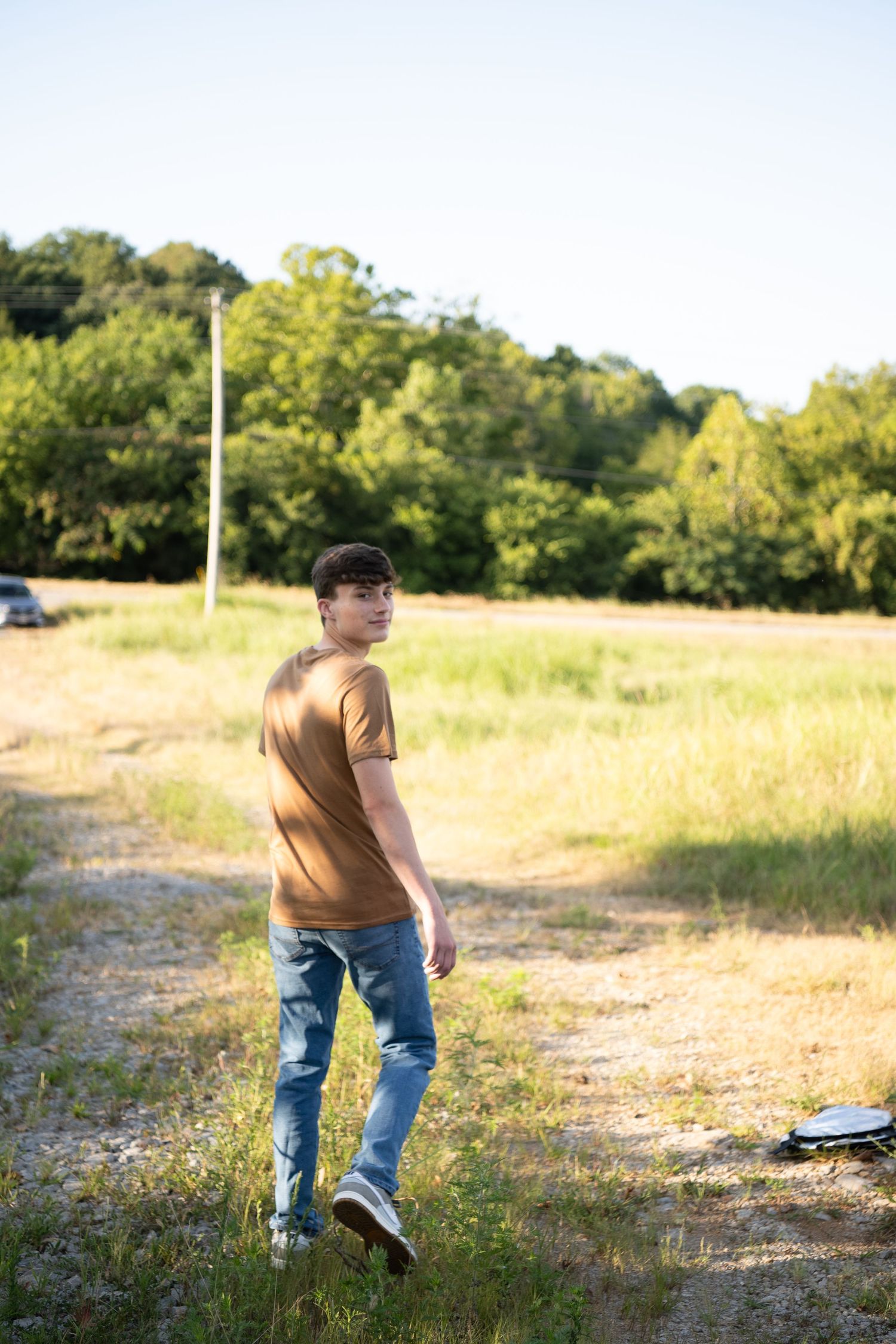 Person in brown shirt and jeans walking on a dirt path through a sunny field with green trees in the background.