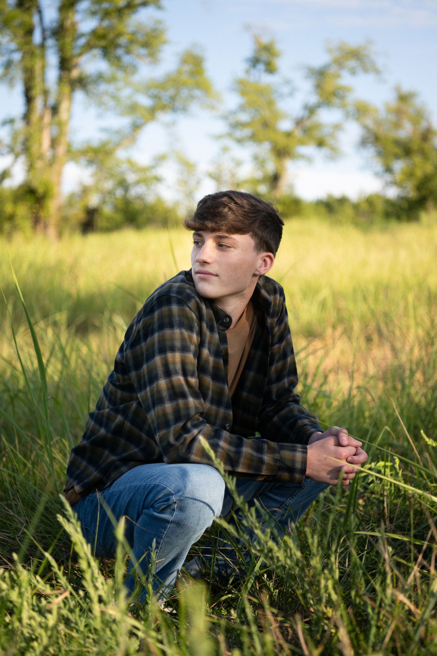 Someone wearing a plaid flannel shirt and jeans sits thoughtfully in tall grass on a sunny afternoon.