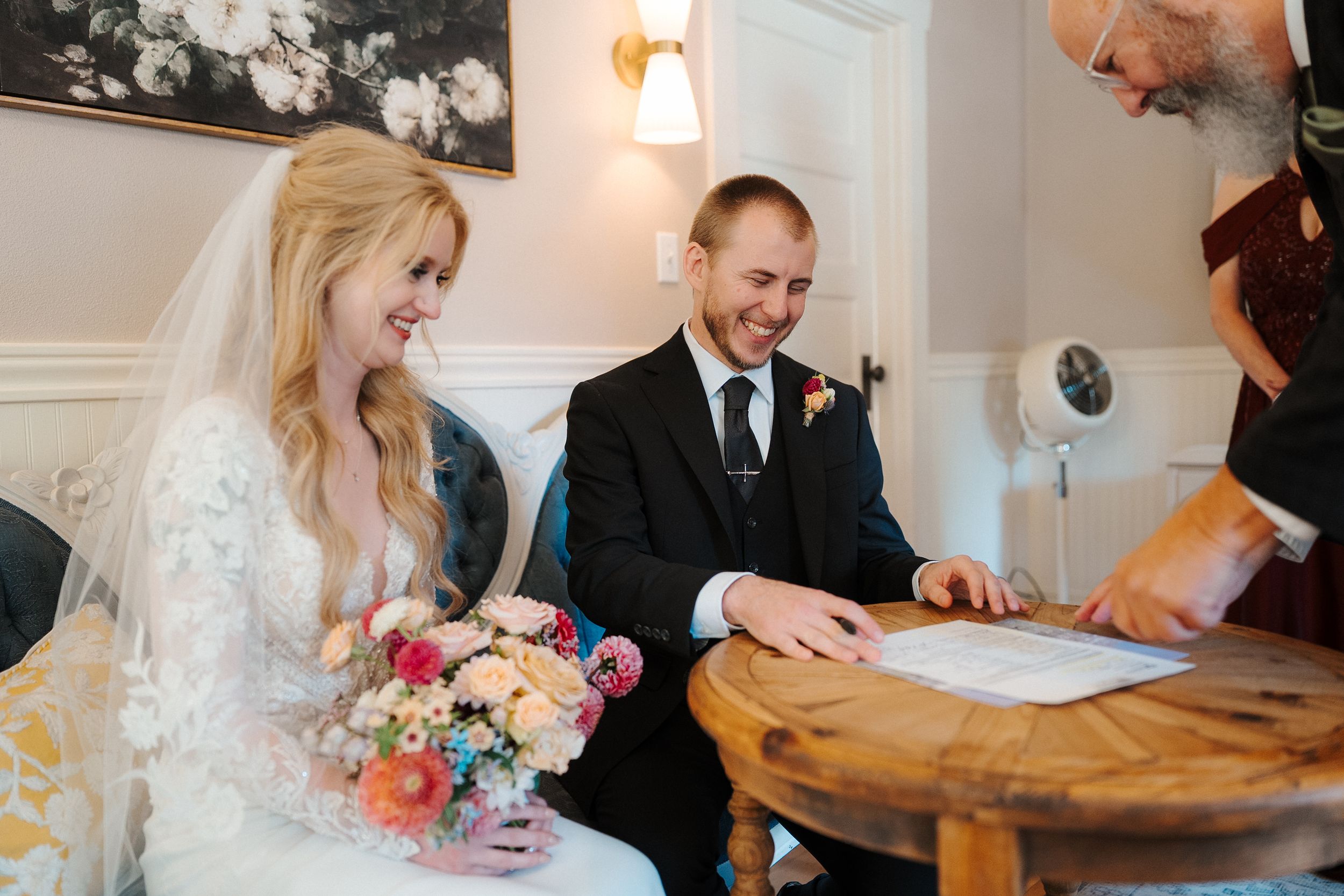 A couple signs wedding documents at a table while guests and witnesses gather around during the ceremony.