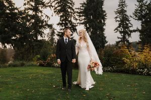 A couple in wedding attire walks together through a scenic garden setting with evergreen trees in the background.
