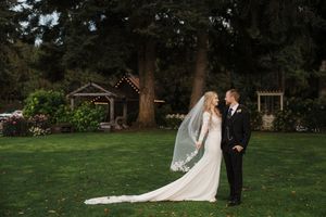 A couple in formal wedding attire embraces on a lush green lawn surrounded by tall trees in a garden setting.