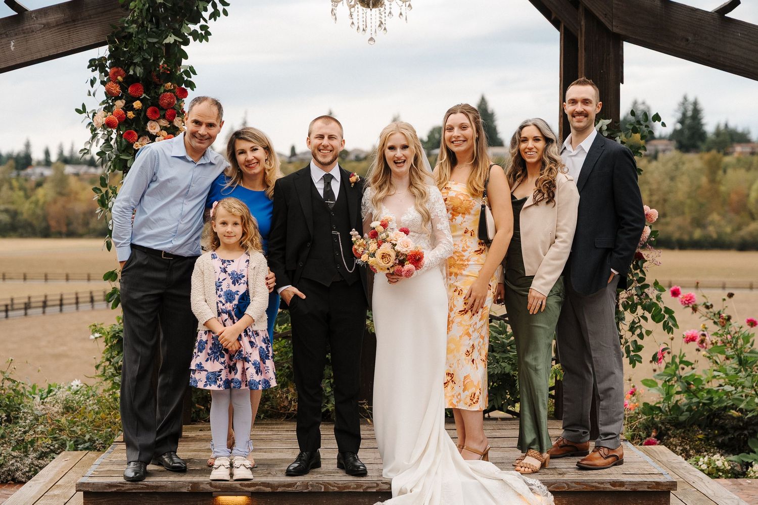 A wedding celebration with guests gathered outdoors at a rustic venue surrounded by beautiful landscaping and flowers.