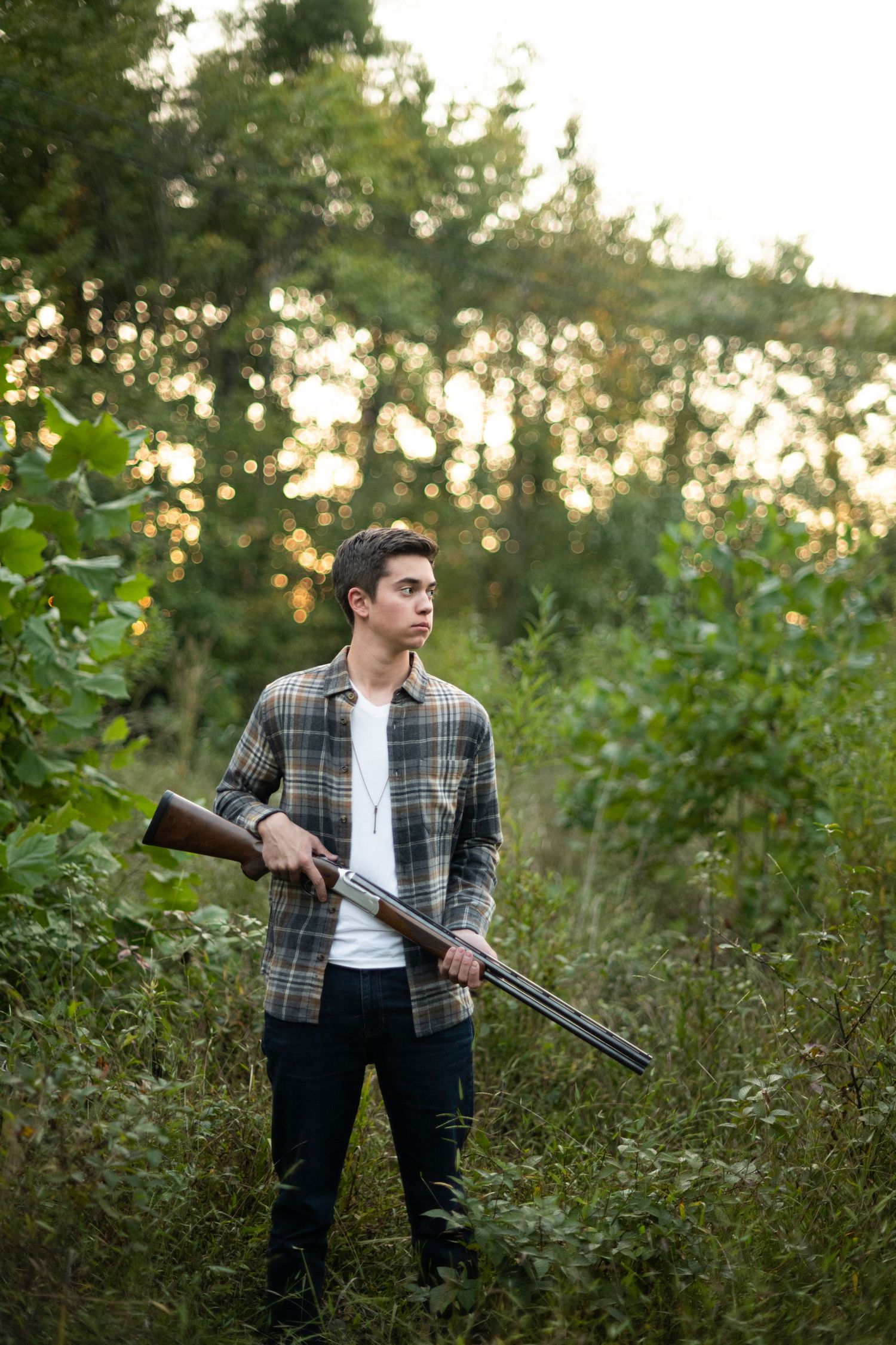 A figure in a plaid shirt and dark pants stands in a lush green field holding a hunting rifle at dusk.