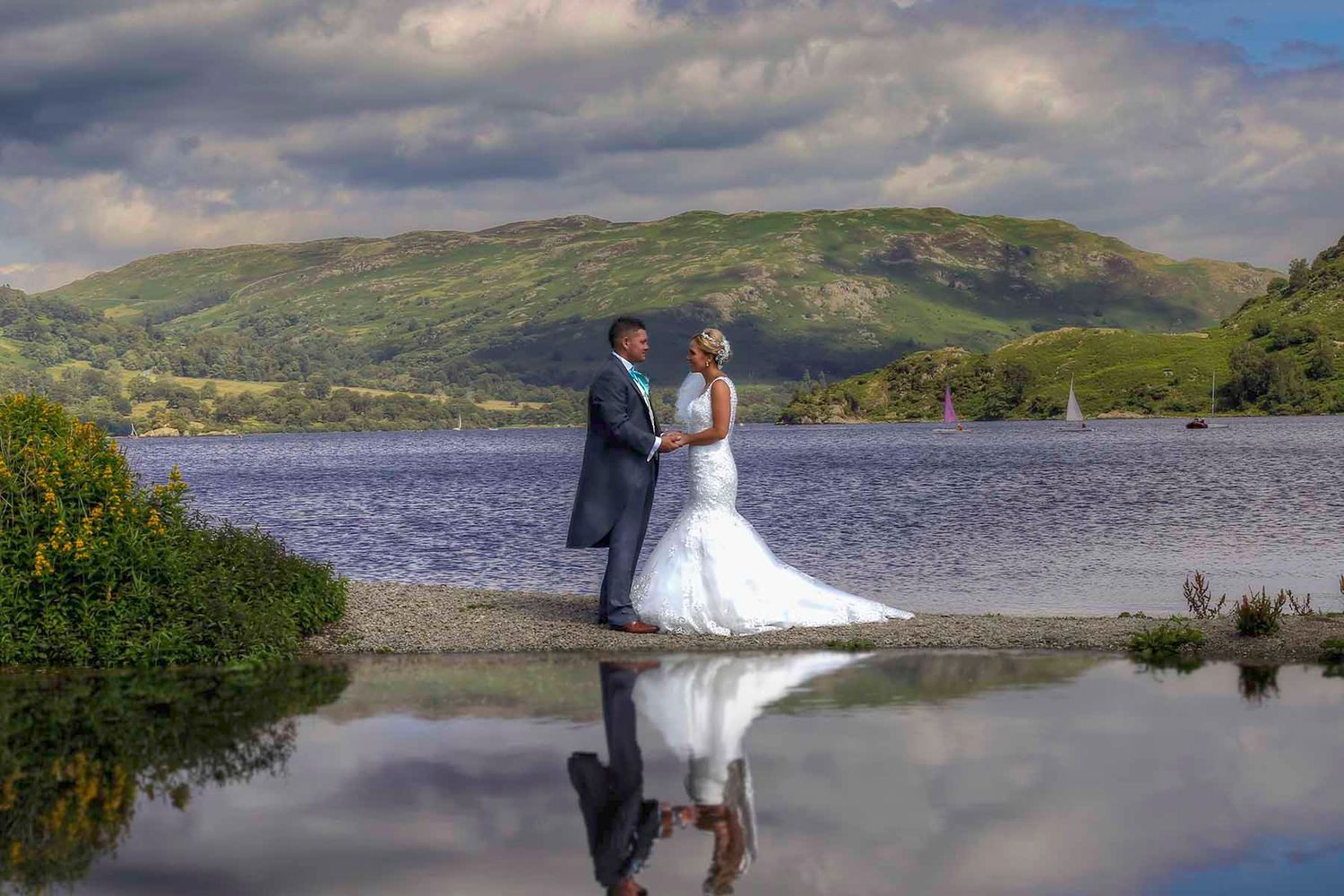 Romantic couple in formal attire standing by scenic lake with mountains and dramatic cloudy sky in background.