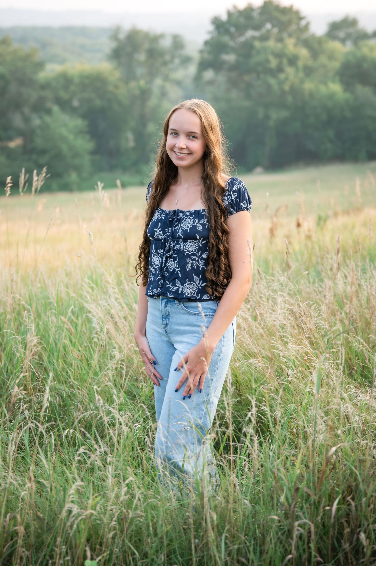 A young woman in a floral navy top sits casually in a grassy field on a summer evening.