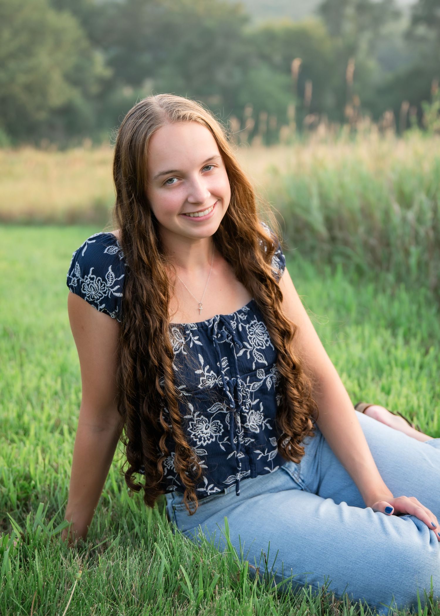 A young woman in a floral top and jeans stands in a sunlit meadow with tall grass and trees in the background.
