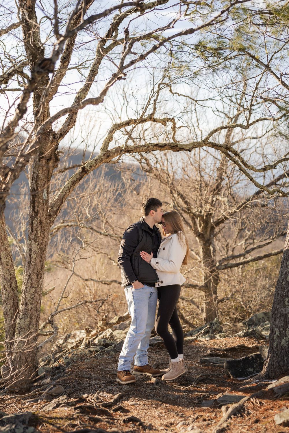 A couple shares an intimate moment on a hilltop surrounded by bare winter trees in natural sunlight.