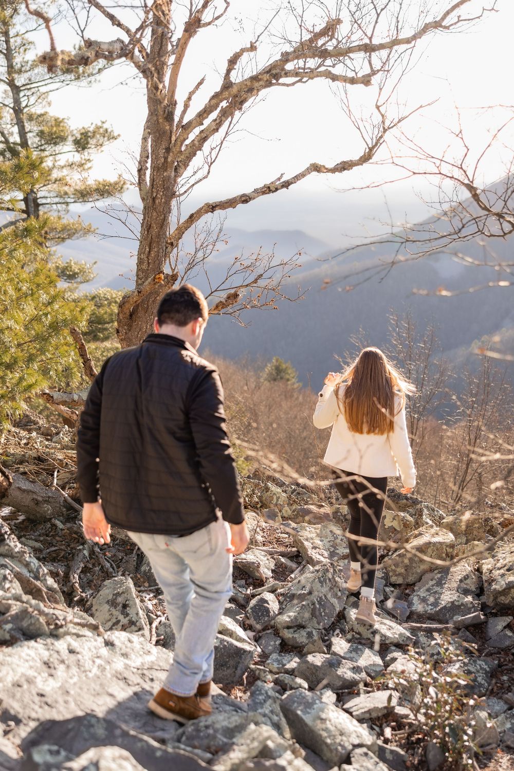 Two people hiking along a rocky mountain ridge with bare winter trees and distant mountain views in the background.