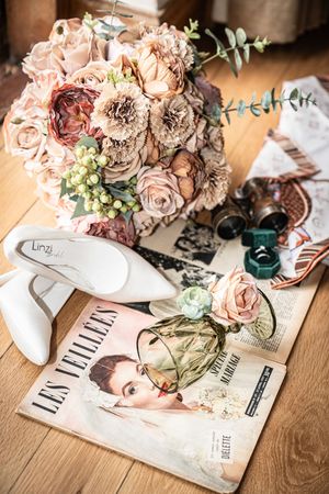 A vintage-style wedding scene with blush roses bouquet, white bridal shoes and retro fashion magazine on wooden floor.