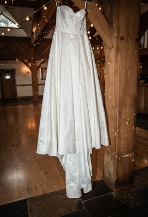 A white wedding dress hangs elegantly from a wooden beam in a rustic barn venue with twinkling string lights.
