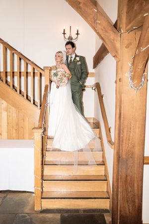 A couple in wedding attire poses on a rustic wooden staircase with exposed beams and a wall-mounted candelabra.