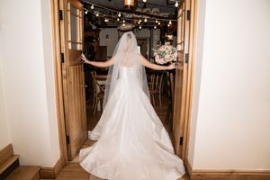 A bride in a flowing white wedding gown and veil twirls in a rustic venue with string lights overhead.