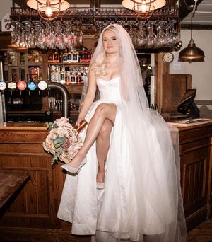 A bride in a white wedding dress poses at a rustic bar with hanging glasses and wooden decor.