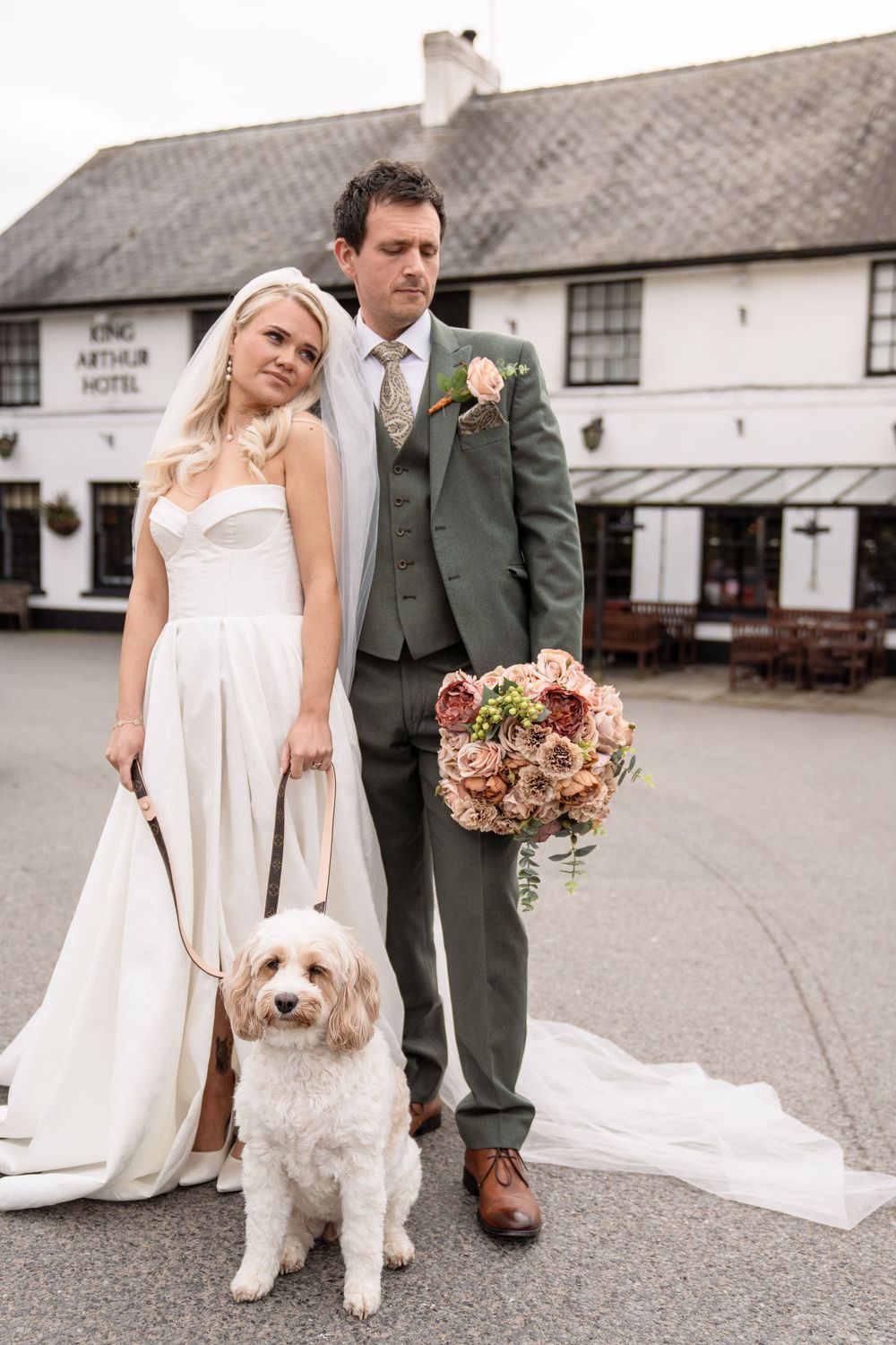A bride and groom pose with their fluffy white dog on their wedding day outside a rustic barn venue.