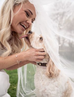 A blonde woman in a white dress shares joyful moments with a fluffy white dog in an outdoor photoshoot.
